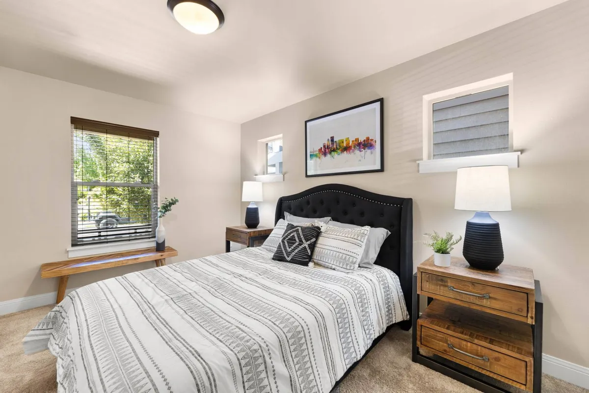 A cozy bedroom awash in natural light, showcasing a comfortable bed, contemporary artwork, and stylish light fixtures. Large windows allow sunlight to filter in, enhancing the sense of serenity and warmth.