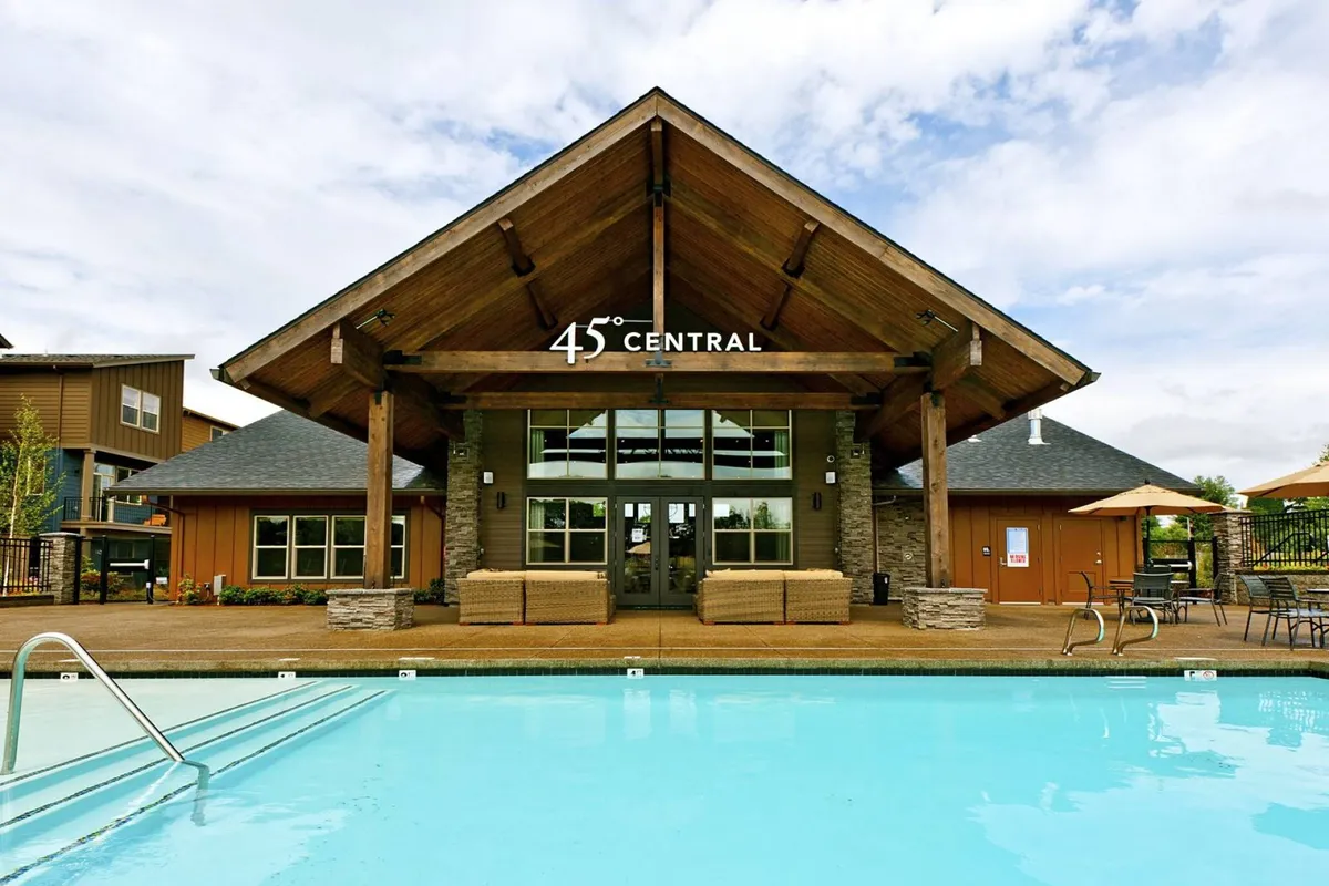 An inviting exterior view of the community pool area, featuring the pool itself, adjacent lounge areas, and the building's main entrance under a distinctive roof structure. The scene captures a welcoming poolside atmosphere, ideal for swimming, sunbathing, and socializing.