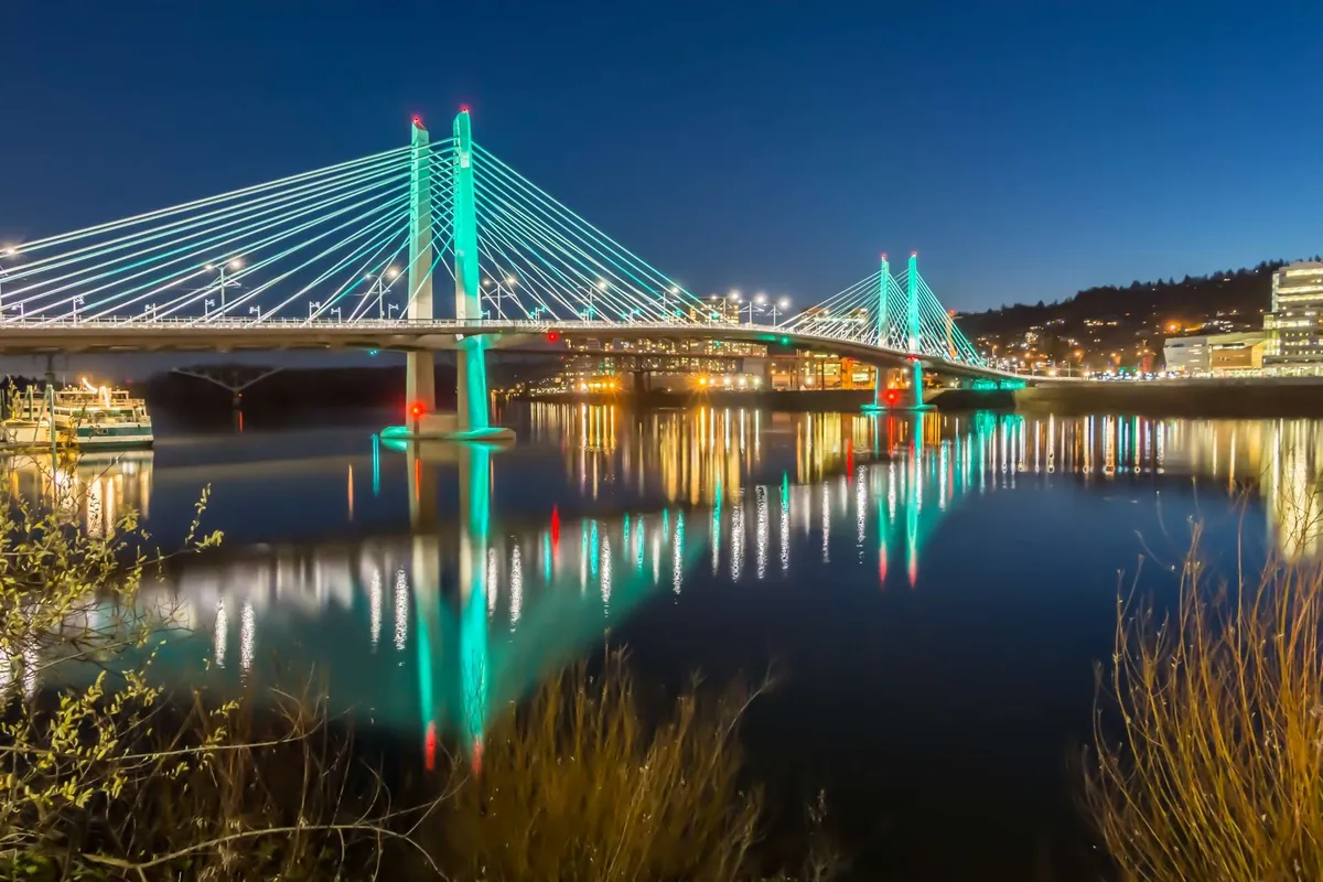 A scenic exterior image featuring a suspension bridge over water, city lights reflecting on the surface, and a lively yet tranquil urban setting. The composition suggests Portland’s vibrant city life and its artistic, photogenic atmosphere.