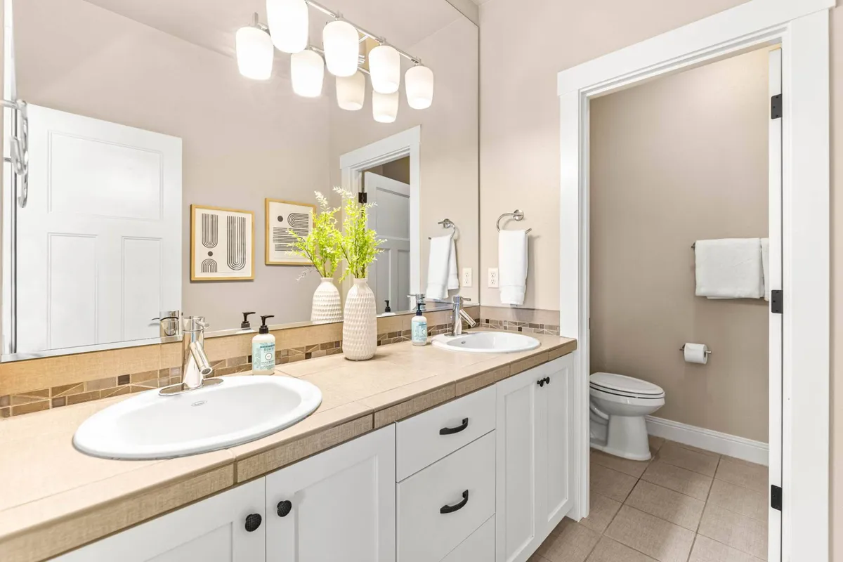 A bright, modern bathroom featuring a double sink vanity, elegant light fixtures, and greenery in a vase. The space is clean, refreshing, and thoughtfully designed for both morning routines and relaxing self-care.
