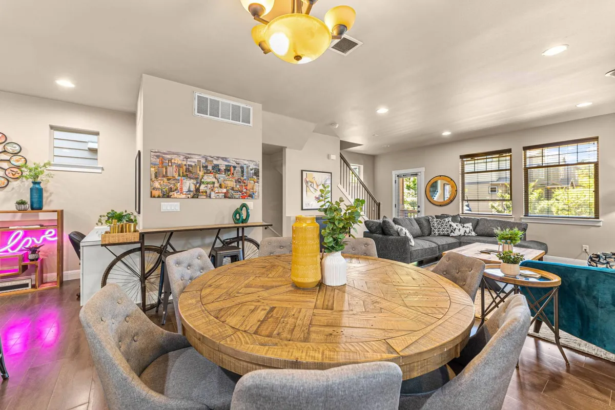 A lively, open-concept space featuring a round wooden dining table, vibrant wall art, a custom neon sign, and a comfortable sofa. The area is sunlit and arranged to encourage group gatherings, conversation, and creativity.