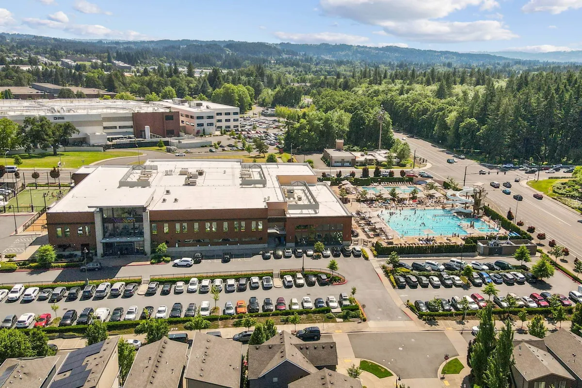 A vibrant exterior shot of the community center complete with a large swimming pool in the foreground, surrounded by greenery and a parking lot. The setting is lively and active, suggesting ample space for recreation, community events, and gatherings.